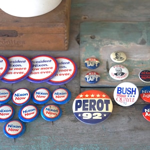 May include: A collection of vintage political campaign buttons featuring various candidates and slogans, including Nixon, Agnew, Perot, Bush, and Quayle. The buttons are red, white, and blue with various designs and text.