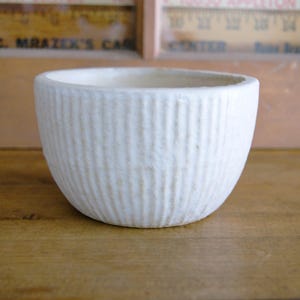 May include: A small, off-white ceramic bowl with a textured, vertically ribbed exterior. The bowl has a rounded shape and sits on a wooden surface. The interior is smooth and unadorned. The bowl is likely for decorative or functional use.