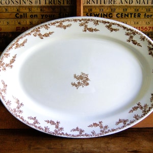 May include: An oval white ceramic serving platter featuring a delicate brown floral pattern along the rim and a small matching floral cluster in the center. The platter is positioned in front of a wooden ruler with visible text.