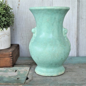 May include: A light green ceramic vase with two handles on the sides. The vase has a rounded body and a flared top.