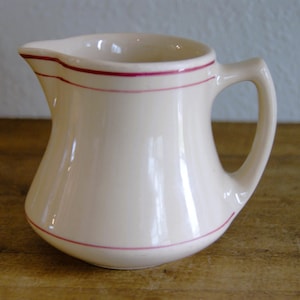 May include: Cream-colored ceramic pitcher with a curved body and a handle. The pitcher is adorned with thin, red-pink stripes near the rim and base. The design is simple and classic, suitable for serving beverages.