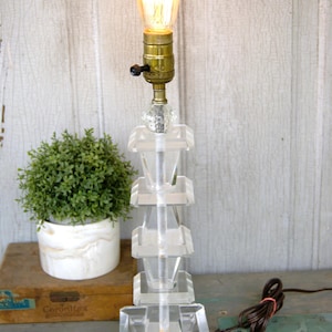 May include: A clear, geometric table lamp with a vintage-style Edison bulb. The lamp has a stacked, rectangular base and a brass bulb socket. A small, green plant in a white pot sits nearby.