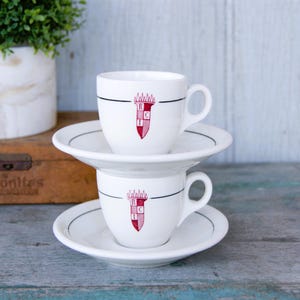 May include: Two white ceramic coffee cups with saucers. The cups have a black line and a red crest with the letters R, C, and I on them.
