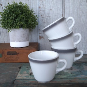 May include: A stack of four white ceramic coffee mugs with a gray rim and handle. One mug is separate, in front of the stack. A small potted plant and a wooden box are in the background.