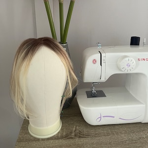 May include: A blonde wig on a white mannequin head with pins in the back. The wig is on a wooden table next to a white sewing machine with the brand name "SINGER" on it. There are pink flowers in a vase behind the mannequin head.