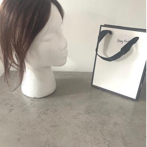 May include: A brown hairpiece sits on a white mannequin head. A white gift bag with black trim and handles is to the right. The bag has a small, round sticker with the text "Envy Hair". The background is a light gray surface and a white wall.
