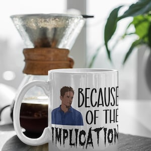 May include: White ceramic mug with the text "BECAUSE OF THE IMPLICATION" and an illustration of a man in a blue shirt. A glass pour-over coffee maker is in the background.