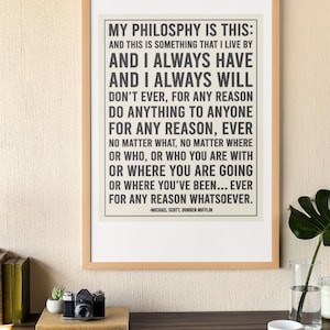 May include: A framed print with the quote "MY PHILOSOPHY IS THIS" by Michael Scott from The Office. The print is in a light wood frame, with a neutral background. A camera, laptop, and coffee cup are on the desk.