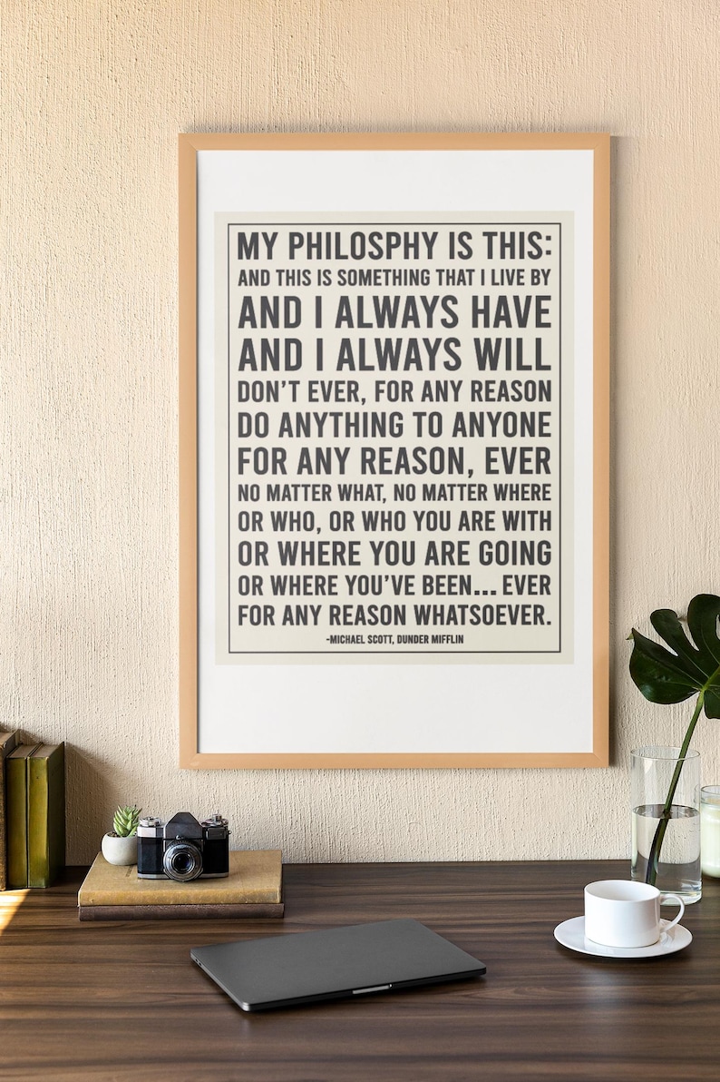 May include: A framed print with the quote "MY PHILOSOPHY IS THIS" by Michael Scott from The Office. The print is in a light wood frame, with a neutral background. A camera, laptop, and coffee cup are on the desk.