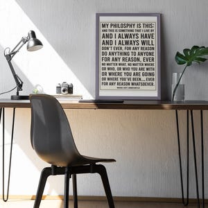 May include: A framed print with the text "MY PHILOSOPHY IS THIS" on a desk with a black chair and lamp. The print has a light gray border and the text is in black. A camera and a plant are also on the desk.