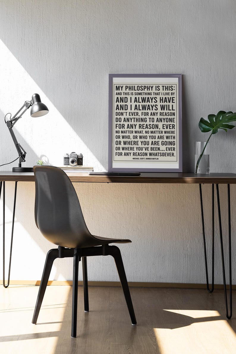 May include: A framed print with the text "MY PHILOSOPHY IS THIS" on a desk with a black chair and lamp. The print has a light gray border and the text is in black. A camera and a plant are also on the desk.
