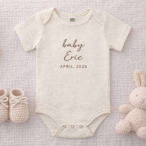 May include: Cream-colored baby onesie with the text "baby Eric APRIL, 2026". The image includes a pair of knitted baby booties, a stuffed bunny, and a sprig of dried grass. The items are arranged on a textured surface.