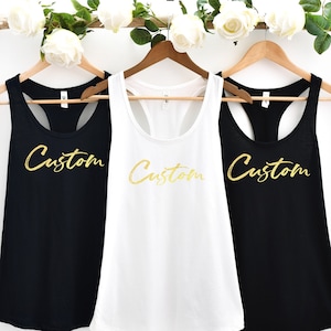 May include: Three racerback tank tops with the word "Custom" written in gold glitter lettering. The tank tops are black, white, and black.
