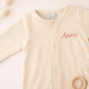 May include: Cream-colored baby onesie with long sleeves and a round neckline. The onesie has the name "Anne" embroidered in pink script on the front. The tag reads "GREEN PEEPS".