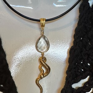 May include: A gold-toned pendant necklace featuring a clear, faceted, pear-shaped gemstone suspended above a textured gold-toned snake charm. The pendant hangs from a simple black cord, showcasing a detailed, scaled serpent design and a polished stone.
