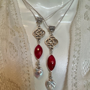 May include: Two silver necklaces with Celtic knot pendants. Each necklace features a faceted, oval, deep red gemstone and a heart-shaped, iridescent pearl. The necklaces hang on delicate silver chains.