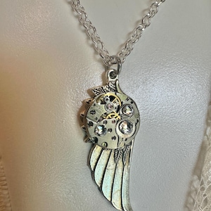 May include: Silver-toned steampunk-style pendant necklace. The pendant features a wing design with intricate gears, crystals, and a small clock face. The necklace has a delicate chain.