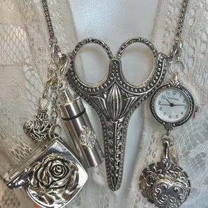 May include: A silver-toned necklace featuring a large pair of scissors as the central pendant. Additional charms include a small pocket watch, a heart-shaped locket, a perfume vial, and a compact with a rose design. The necklace is displayed against a white lace background.