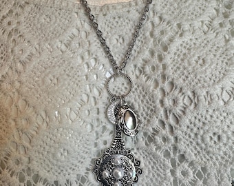 May include: A silver-toned pendant necklace featuring a long chain, a crescent moon charm, an oval locket, and a large ornate filigree centerpiece adorned with four white pearls and a single clear teardrop crystal dangling at the bottom.