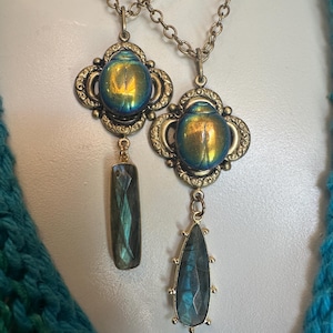 May include: Two necklaces featuring ornate brass settings with iridescent blue and gold beetle-shaped cabochons. One pendant includes a rectangular faceted gray stone drop, while the other features a teardrop-shaped faceted gray stone pendant.