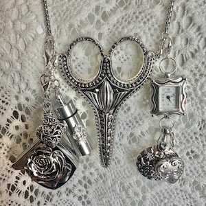 May include: A silver-toned necklace featuring a large central scissor pendant. Attached to the chain are several charms, including a square watch with the text Narmi, a rose, a heart, and a small cylindrical container, all with intricate metal detailing.