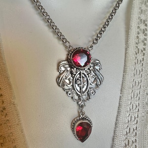 May include: Silver-toned necklace featuring a pendant with a large, round, red gemstone and a smaller, teardrop-shaped red gemstone. The pendant has an ornate, filigree design. The chain is a simple link style.