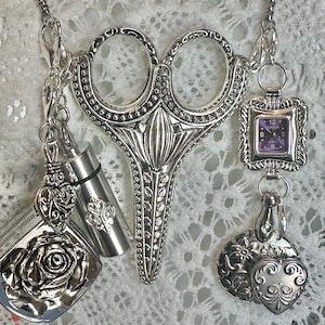 May include: A silver-toned necklace featuring a large central scissor-shaped pendant, a small square watch with a purple face labeled Geneva, and various dangling charms including a rose, hearts, and a cylindrical metal container on a chain.