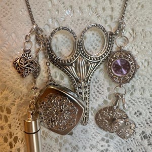 May include: A silver-toned necklace featuring a large central scissor pendant, a small round watch with a purple face, a heart-shaped charm, a square filigree locket, a cylindrical metal vial, and a round floral embossed charm.