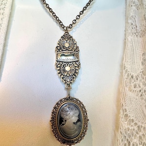 May include: A bronze-toned necklace featuring a chain attached to an ornate, elongated metal pendant with a keyhole cutout. Below this hangs an oval-shaped cameo pendant with a dark background and a light-colored profile portrait of a person.