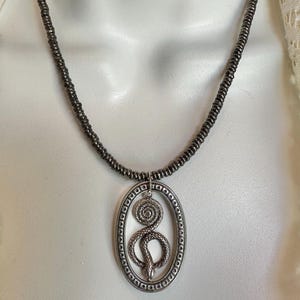 May include: A necklace featuring a dark metallic beaded chain and an oval pendant. The pendant contains a coiled snake design within a textured border. The metal has a gunmetal gray finish, and the snake is depicted in a curled, resting position.