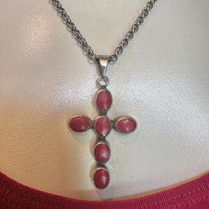 May include: A silver chain necklace featuring a cross-shaped pendant. The pendant is set with five smooth, oval-shaped red stones arranged in a cross pattern. The metal has a polished finish, and the chain consists of interconnected circular links.
