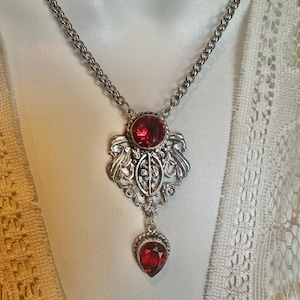 May include: Silver-toned necklace featuring a pendant with a large, round, red gemstone at the top and a teardrop-shaped red gemstone at the bottom. The pendant has an ornate, filigree design. The necklace is displayed on a white surface.