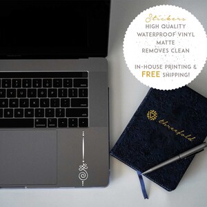 May include: A white laptop with a silver keyboard and a white sticker on the trackpad. The sticker is a simple design with a spiral and dots. A blue notebook with gold lettering that says "threefold" is on a white surface next to the laptop. A silver pen is on top of the notebook.