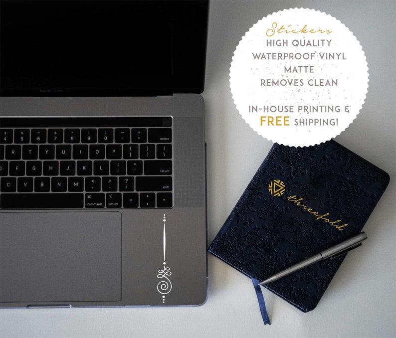 May include: A white laptop with a silver keyboard and a white sticker on the trackpad. The sticker is a simple design with a spiral and dots. A blue notebook with gold lettering that says "threefold" is on a white surface next to the laptop. A silver pen is on top of the notebook.