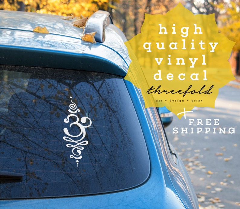 May include: White vinyl decal of an Om symbol on a blue car with the text "high quality vinyl decal threefold art + design + print FREE SHIPPING" in yellow.