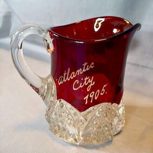 Puede incluir: Una jarra de cristal rojo rubí con un asa transparente y una base decorativa de cristal tallado. Las palabras "Atlantic City 1905" están inscritas en escritura dorada en el lateral. La jarra tiene un borde acampanado.