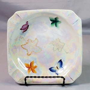 May include: A square, white ceramic dish with a iridescent glaze. The dish is decorated with hand-painted butterflies, leaves, and flowers in shades of orange, yellow, green, blue, and purple.