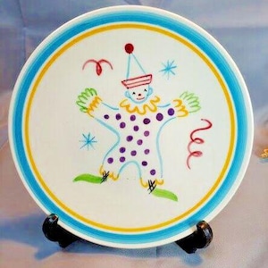 Puede incluir: Un plato de cerámica blanco con un diseño de payaso caprichoso. El payaso es azul claro con lunares morados, manos y pies verdes y un sombrero rojo. El plato tiene un borde azul y amarillo y se muestra en un soporte negro.