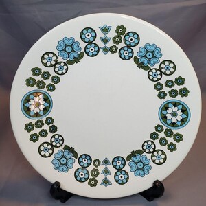 May include: White ceramic dinner plate with a blue and green floral pattern around the edge. The pattern features stylized flowers, leaves, and circles.