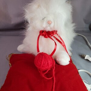 May include: White plush cat toy with a red bow and a red yarn ball sitting on a red velvet pillow with gold trim.