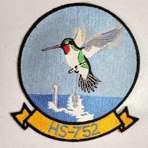 Könnte beinhalten: Gestickter Aufnäher mit einem Kolibri im Flug über einer blauen Meereslandschaft. Der Vogel hat grüne, rote und weiße Details. Der Aufnäher enthält den Text "HS-752" auf einem gelben Banner und einen schwarzen Rand.