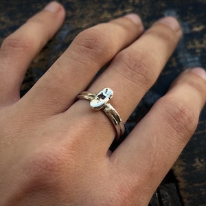 Könnte beinhalten: Ein silberfarbener Ring mit einer dünnen, glatten Ringschiene und einem zentralen, länglich ovalen Element, das eine texturierte Oberfläche mit dunklen Flecken aufweist. Das Metall ist poliert und das Design besteht aus einem einzelnen Ring.