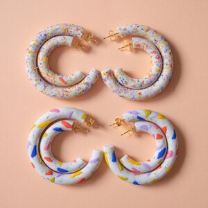 Könnte beinhalten: Vier Paar weiße Creolen mit bunten Sprenkeln in Gelb, Blau, Rosa und Lila. Die Ohrringe haben goldfarbene Stifte und sind auf einem hellpfirsichfarbenen Hintergrund angeordnet. Ein lustiges und verspieltes Accessoire.