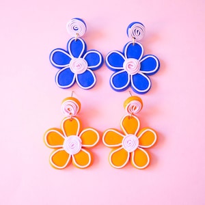Op de afbeelding: Een paar blauwe en een paar oranje bloemvormige oorhangers. De oorhangers hebben een wit midden en een wit swirl-detail.