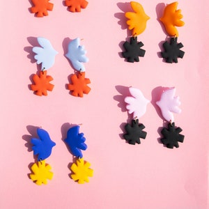 May include: Assortment of colourful dangle earrings featuring a bird shape with a flower-like design. The earrings are available in blue, orange, light blue, pink and yellow.