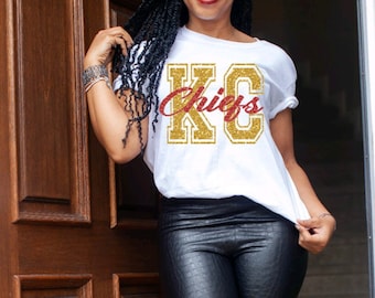 May include: A white t-shirt with a gold glitter "KC Chiefs" graphic. The shirt is being worn by a person with long black hair and black pants.