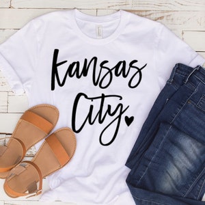Op de afbeelding: Wit T-shirt met de woorden "Kansas City" in zwart script, een klein hartje en opgerolde mouwen. Het shirt wordt gecombineerd met een blauwe jeans en bruine sandalen.