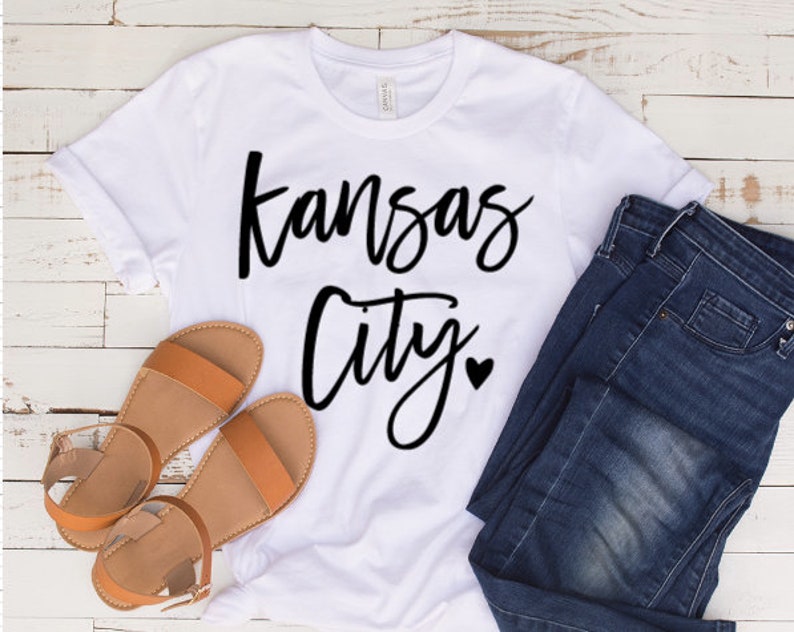 Op de afbeelding: Wit T-shirt met de woorden "Kansas City" in zwart script, een klein hartje en opgerolde mouwen. Het shirt wordt gecombineerd met een blauwe jeans en bruine sandalen.