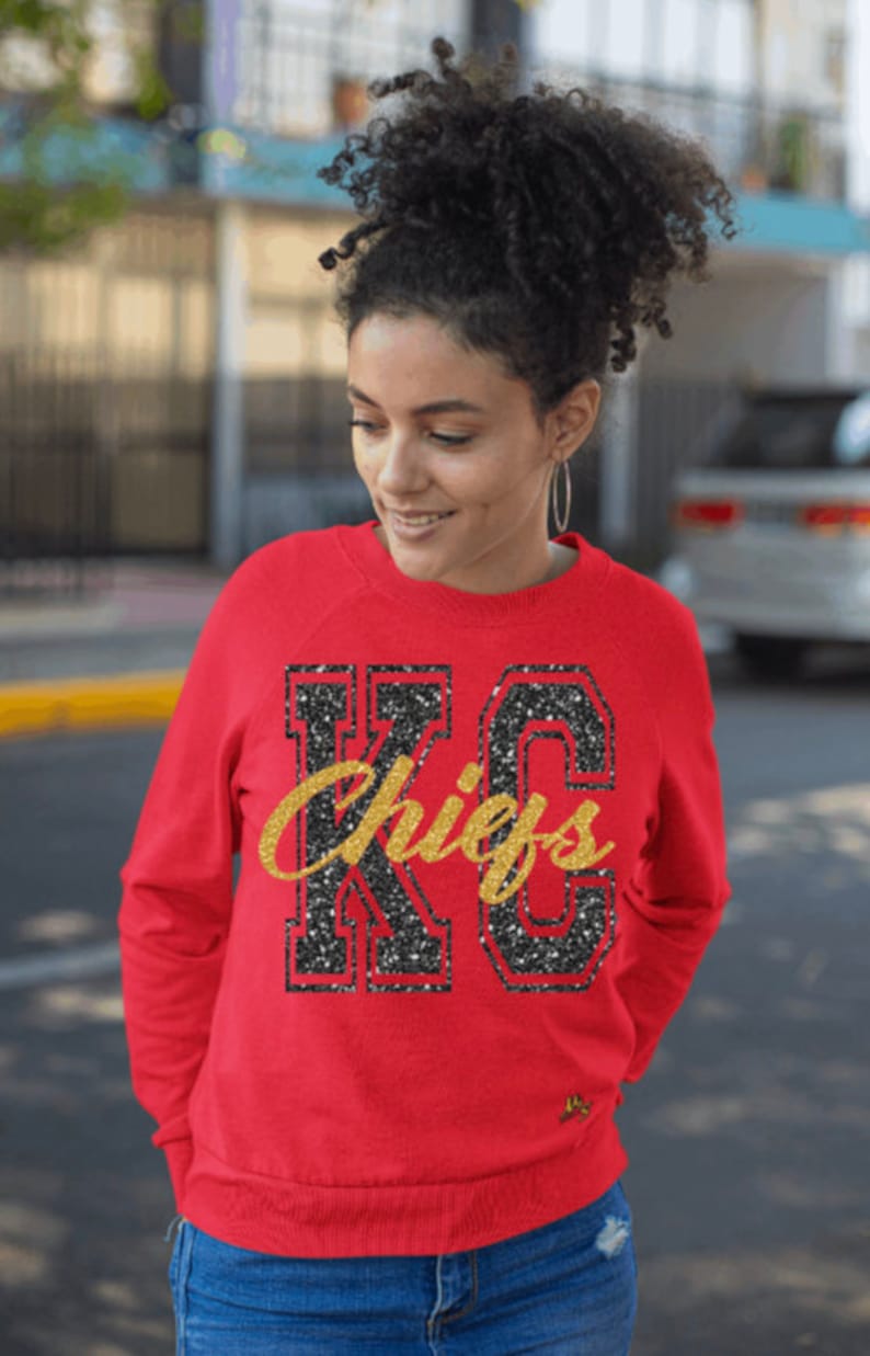 Op de afbeelding: Rood sweatshirt met een zwart en gouden glitterontwerp met de tekst "KC Chiefs".