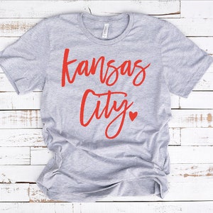 Op de afbeelding: Een lichtgrijs t-shirt met de tekst "Kansas City" in rode letters. Onder het woord "City" staat een klein rood hartje. Het shirt is gemaakt van een zacht materiaal en heeft korte mouwen.